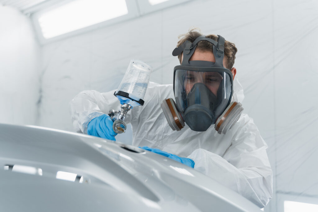 Car painter in protective clothes and mask painting and varnish automobile bumper in chamber